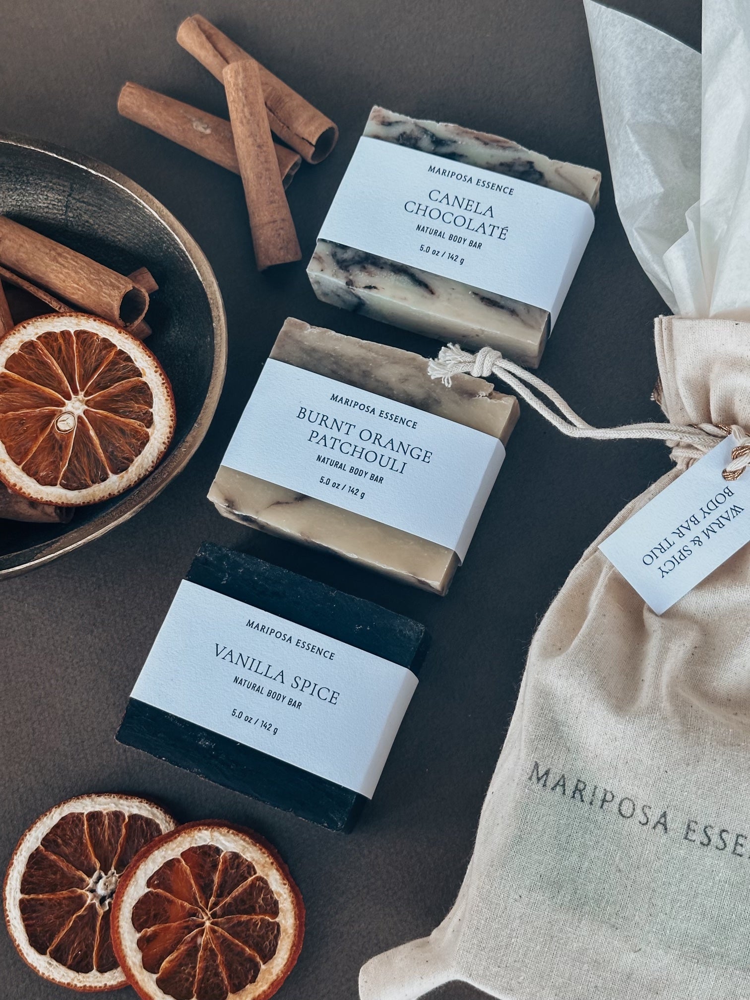 Warm & Spicy Body Bar Trio in a muslin bag with tissue displayed with a bowl of cinnamon sticks and dehydrated oranges